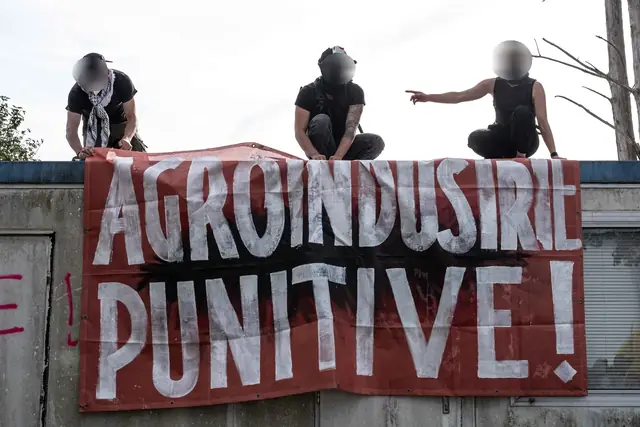 Three masked people on a roof above a red banner: “Punitive agro-industry!” (“Agro-industrie punitive !”)