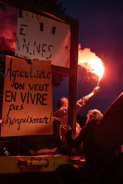 At night, a red flare lights up the sign “Farming: we want to make a living from it, not poison ourselves!” (“Agriculture : on veut en vivre, pas s’empoisonner !”)
