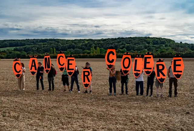 Protesters lined up in a field spell out the words “Cancer Anger” (“Cancer colère”) with large orange letters
