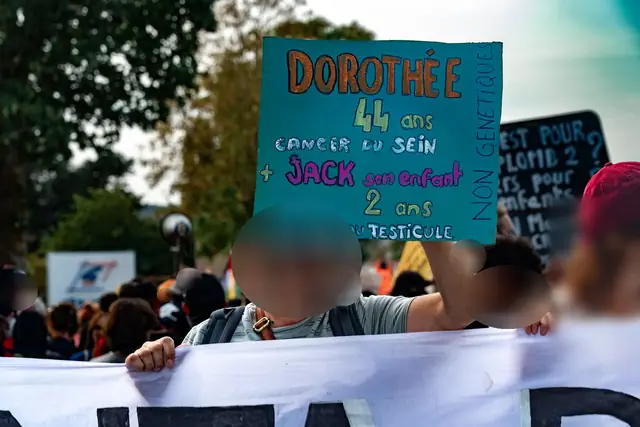 A protester holds a sign: “Dorothée, 44, breast cancer, and Jack, her child, 2, testicular cancer” (“Dorothée, 44 ans, cancer du sein, et Jack, son enfant, 2 ans, cancer du testicule”)