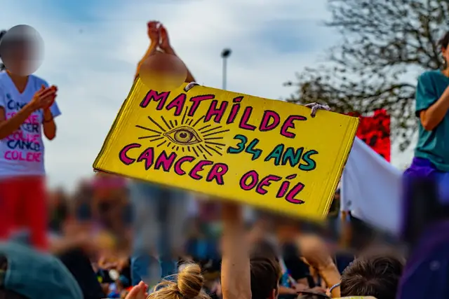 A yellow sign, “Mathilde, 34, eye cancer” (“Mathilde, 34 ans, cancer de l’œil”), held up above the crowd